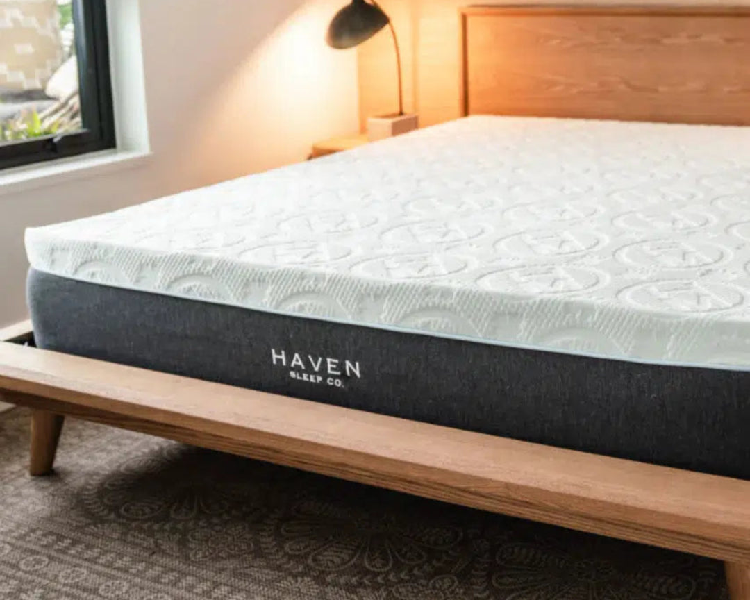 A Haven Sleep Co. mattress from the Side Sleeper Bundle is elegantly displayed on a wooden platform bed in a modern, minimalist bedroom. The mattress, with its cool-to-the-touch bamboo cover and advanced pressure relief features, is designed specifically for side sleepers. The room's serene ambiance, enhanced by a softly lit bedside lamp and neutral decor, emphasizes the luxurious comfort and support offered by the LUX Hybrid Pillowtop or LUX Rejuvenate mattress. This mattress provides optimal support for h
