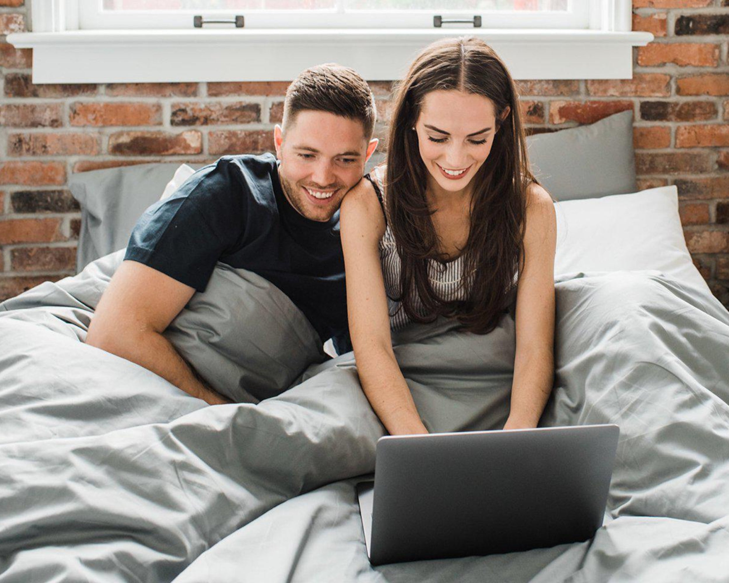 Man and woman cozied up in Haven Mattress bed-in-box bed, tucked into Storm Grey Bedface sheets and duvet cover. The couple is both looking at a grey laptop while the woman is typing