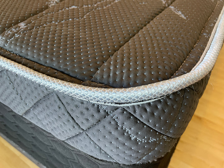 Close-up of the corner edge of the Haven HD Hybrid mattress, highlighting the intricate stitching and textured black and gray fabric. Designed for plus-size sleepers, this mattress offers firm and extra-firm support with high-density materials and Talalay latex. The detailed view emphasizes the premium craftsmanship and durability of the mattress, ensuring long-lasting comfort.