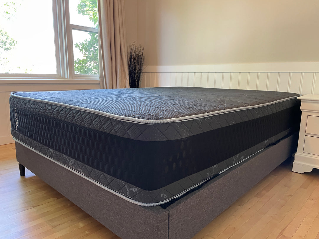 Angled view of the Haven HD Hybrid mattress set up in a well-lit bedroom with natural light from a nearby window. The mattress, designed for plus-size sleepers, features a black and gray honeycomb pattern with a firm and extra-firm support system. The elegant design and high-density materials provide a comfortable and durable sleep surface, perfect for those needing additional support.