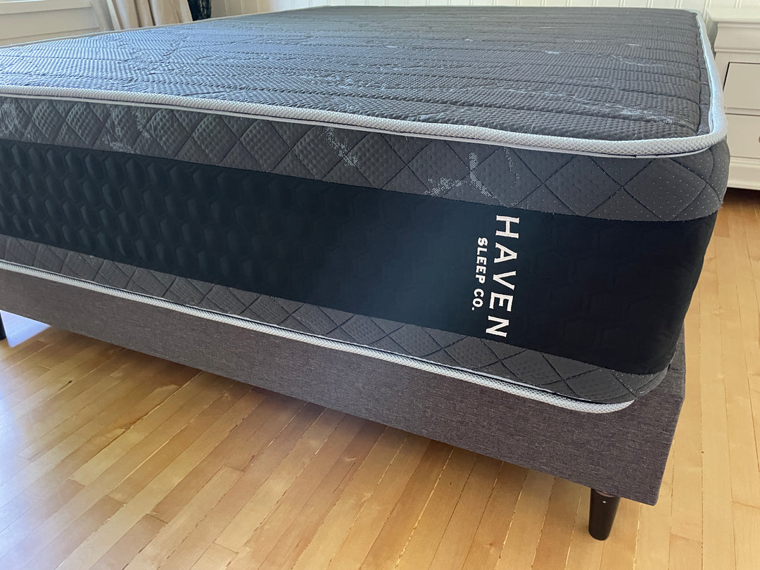 Side view of the Haven HD Hybrid mattress, emphasizing the black and gray honeycomb design with visible Haven Sleep Co. branding. Positioned in a bedroom with a modern bed frame, this mattress is designed for plus-size sleepers, featuring 7-zone HD coils and Talalay latex for firm and extra-firm support. The image highlights the high-quality materials and construction for enhanced durability and comfort.