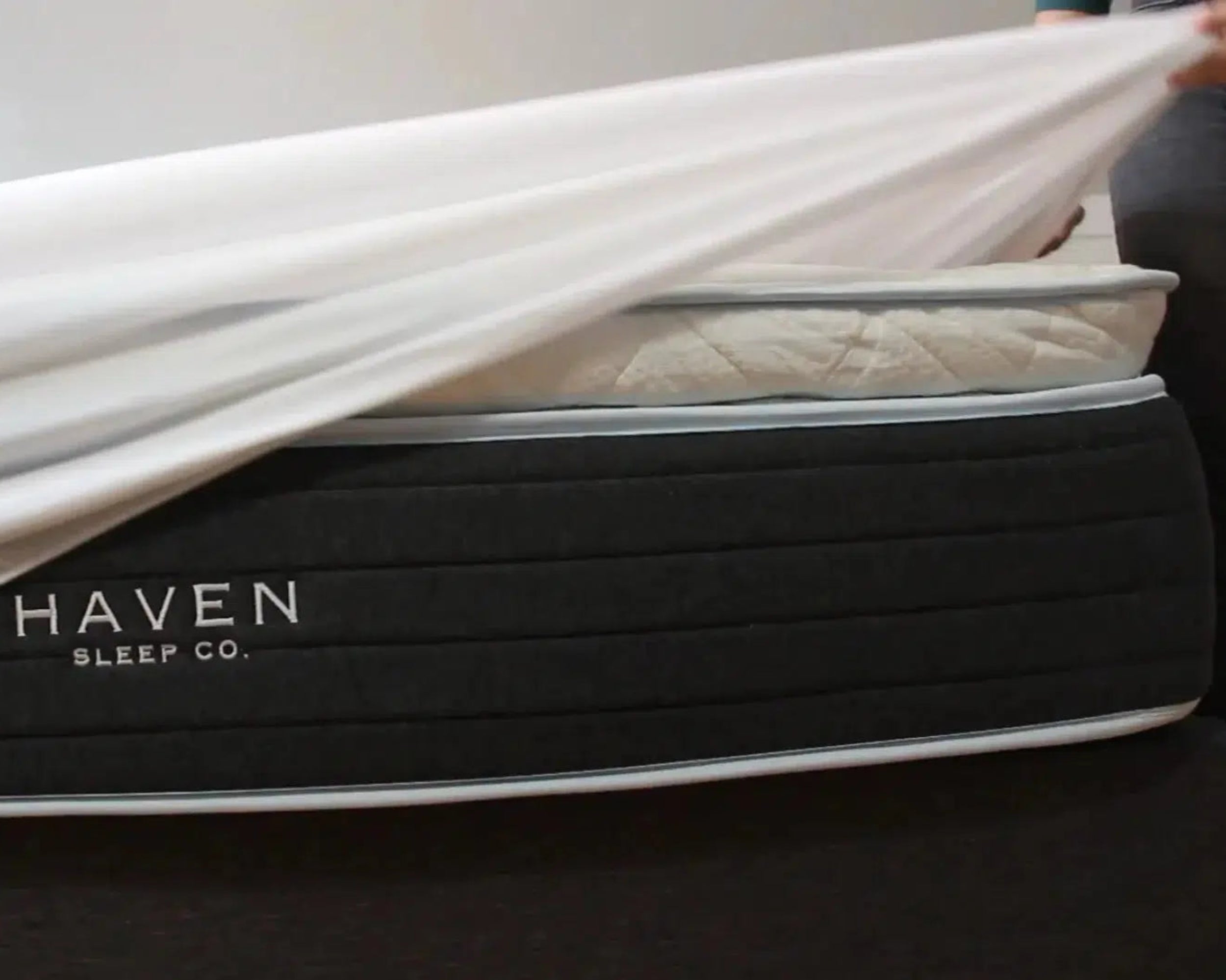 Person placing a smooth and lightweight mattress protector on a Haven Sleep Co. mattress, highlighting its waterproof, hypoallergenic, and breathable features. The protector has deep pockets and is made with some natural bamboo content, offering antimicrobial and allergen-proof protection, made in Canada.