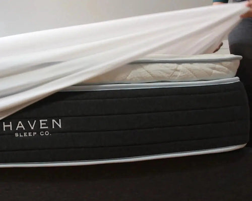 Person placing a smooth and lightweight mattress protector on a Haven Sleep Co. mattress, highlighting its waterproof, hypoallergenic, and breathable features. The protector has deep pockets and is made with some natural bamboo content, offering antimicrobial and allergen-proof protection, made in Canada.