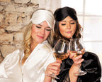 Two women in luxurious satin sleepwear, one in white and the other in black, enjoy a glass of wine while wearing satin eye masks from the Dream Satin Sleep Set. The set includes a satin pillowcase, scrunchie, and eye mask, offering a cruelty-free and sustainable alternative to traditional silk, ensuring frizz-free hair and a rejuvenating sleep.
