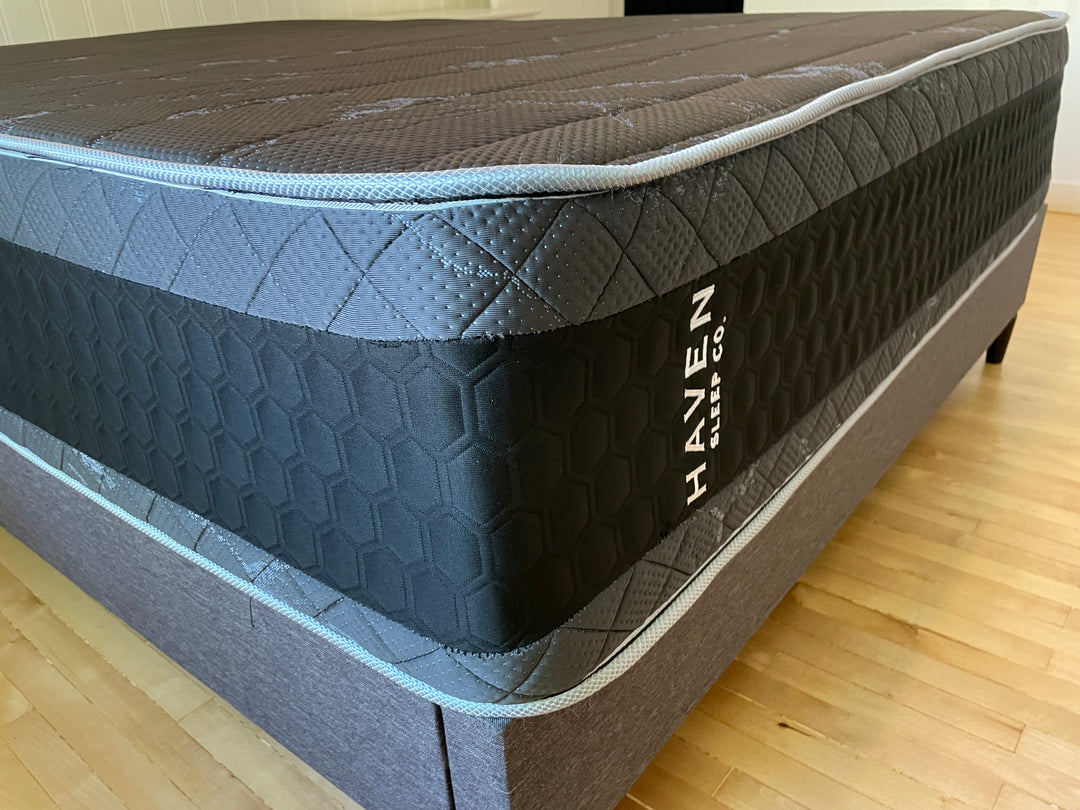 Angled close-up view of the Haven HD Hybrid mattress, highlighting the black and gray honeycomb design and visible Haven Sleep Co. branding. The mattress, designed for plus-size sleepers, features high-density materials and advanced support systems, including 7-zone HD coils and Talalay latex. The image emphasizes the mattress's sturdy construction and premium comfort, ideal for those seeking firm and extra-firm support.