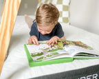 Child reading a book on a mattress with a wooden crib frame in the background
