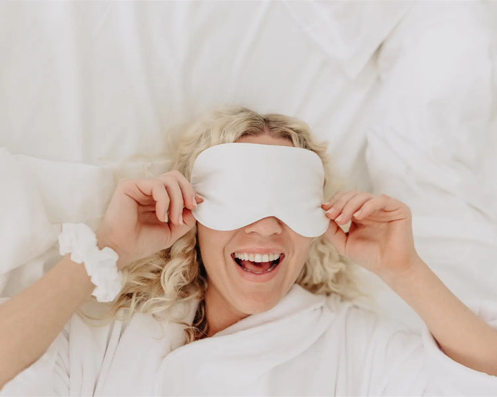 Woman wearing a white sleep mask in bed, smiling.