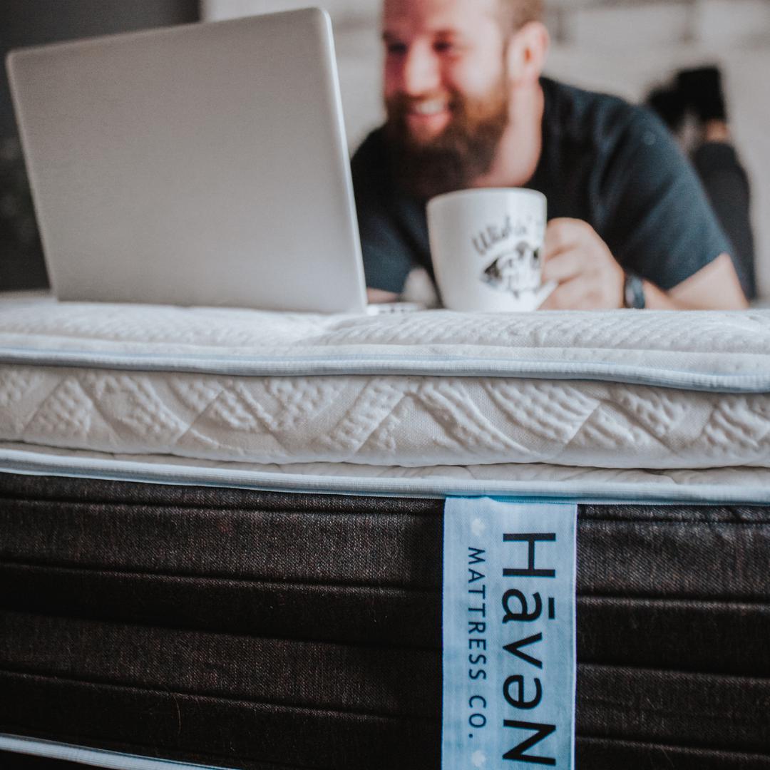 Homme allongé sur un matelas Haven, tenant une tasse de café et regardant un ordinateur portable blanc avec l'étiquette du matelas Haven sur le devant.