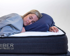 Person sleeping on a mattress with a pillow, wearing a blanket, on a white background. Hiber Mattress Co branding visible.