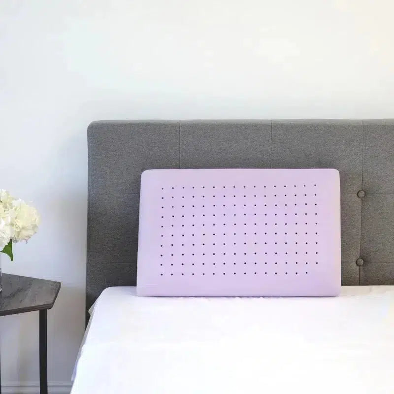 Lavender Infused Gel Foam Pillow propped up against a gray upholstered headboard, showcasing its lavender color and ventilation holes, designed for cooling and pressure relief.