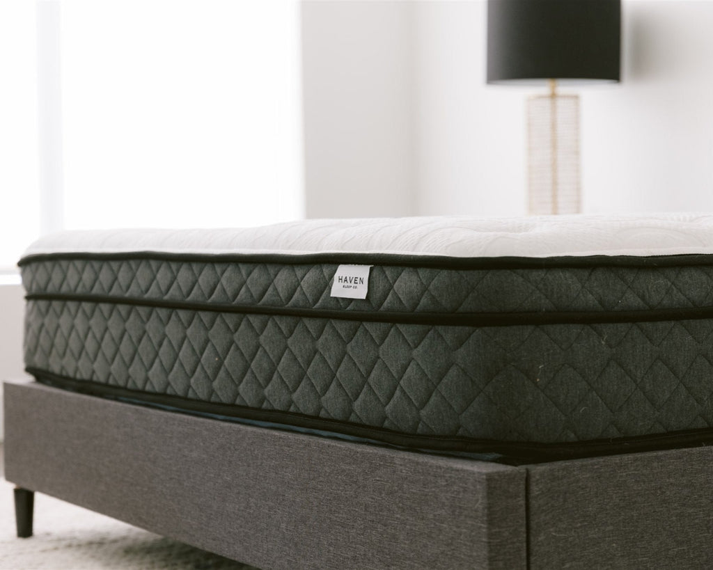 Side view of the Haven Pocket Coil Mattress with a white quilted top and dark grey diamond-patterned sides, positioned on a grey upholstered bed frame in a modern bedroom. The mattress label displays the Haven Sleep Co. logo, emphasizing its premium construction, including 884 individually wrapped coils and a bamboo cover with cool touch technology.