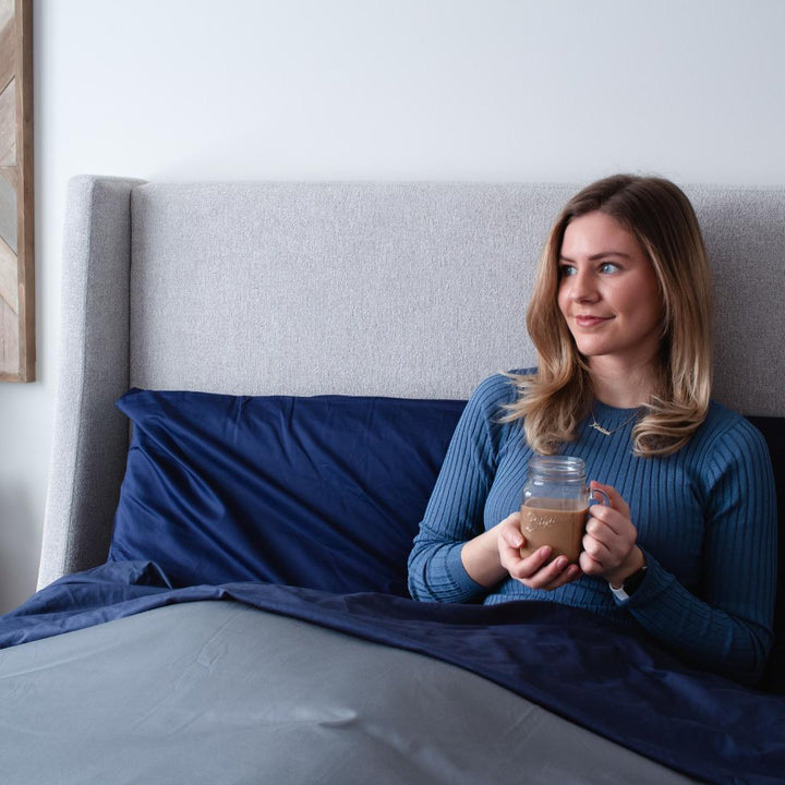 Femme assise dans son lit avec une tasse de café et la collection Bedface sateen de haven sleep company en marine et gris